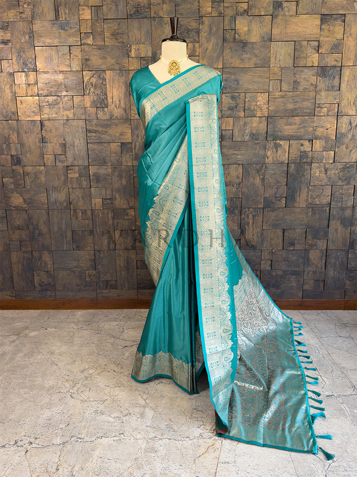 Teal saree with a patterned border on a mannequin against a wooden paneled wall. Full front display of a solid teal green satin silk saree featuring a heavy woven zari border draped gracefully on a mannequin.