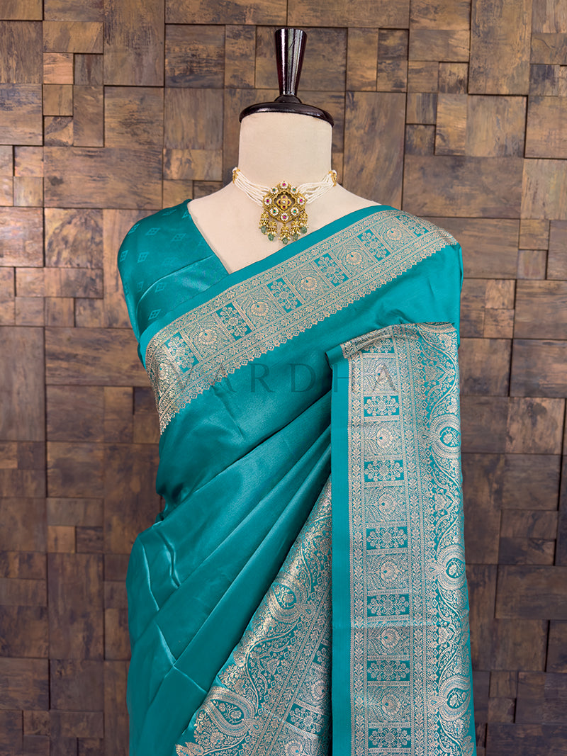 Teal saree with intricate patterns on a mannequin against a wooden paneled wall. Close-up of a sea green woven satin ethnic saree styled on a mannequin display, paired with a traditional pearl choker necklace.