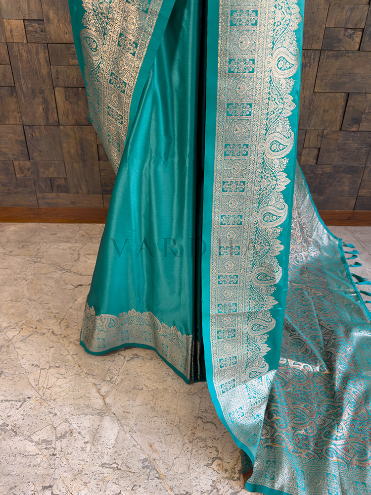 Teal saree with intricate patterns on a wooden floor Lower pleats and fluid drape of a glossy teal Banarasi-style satin silk saree showcasing the broad geometric floral border.