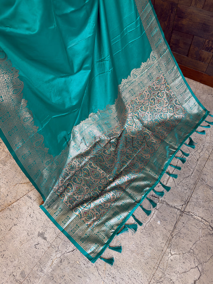 Teal saree with intricate patterns on a stone floor Detailed floor view of the rich, densely woven zari pallu with matching tassels on a vibrant teal green satin saree.