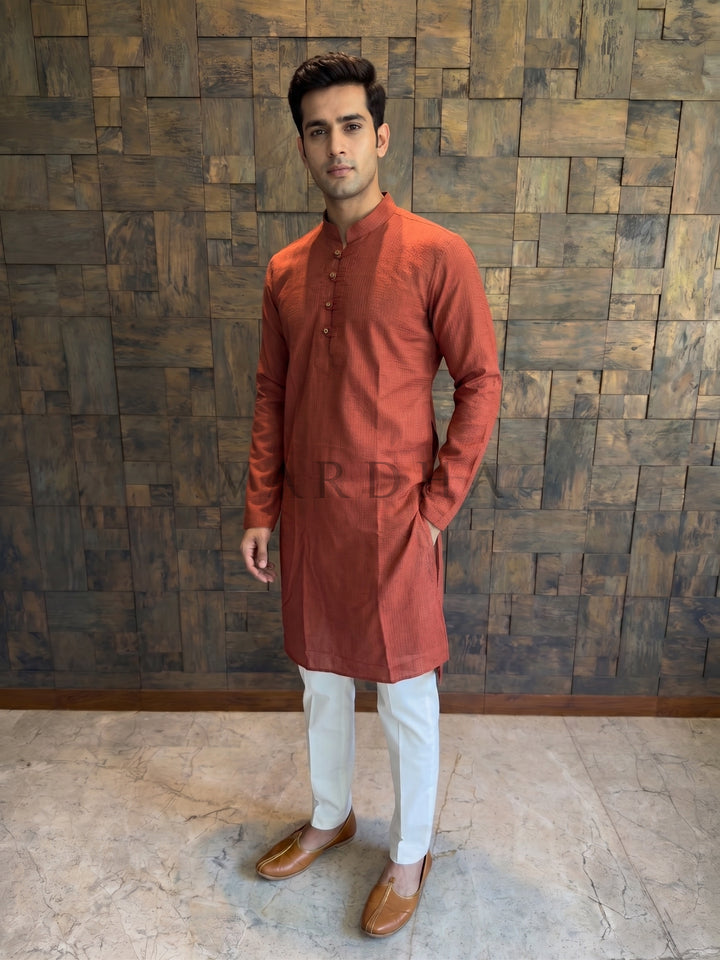 Man wearing a rust-colored kurta with white pants against a wooden paneled wall. Terracotta orange cotton kurta for men featuring a woven micro-check texture and an extended chest placket with traditional fabric button loops.
