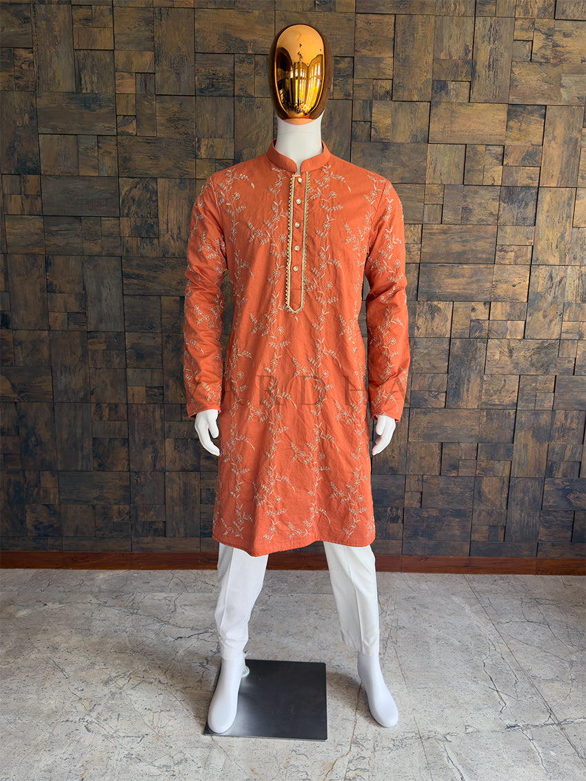 Mannequin wearing an orange kurta with white patterns against a wooden paneled wall. Front view of a man wearing a vibrant terracotta orange viscose kurta featuring all-over floral vine embroidery.