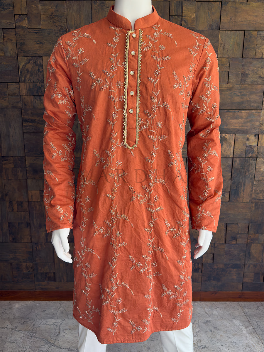 Orange kurta with intricate embroidery on a wooden paneled background. Close-up of the terracotta orange kurta showcasing the intricate floral threadwork and a gold zig-zag bordered placket. Tailored with a clean Mandarin collar. The embellished placket is fastened with premium, crystal-studded designer buttons, adding an extra layer of festive luxury. A classic knee-length Long Kurta with full, straight sleeves and a straight hem.