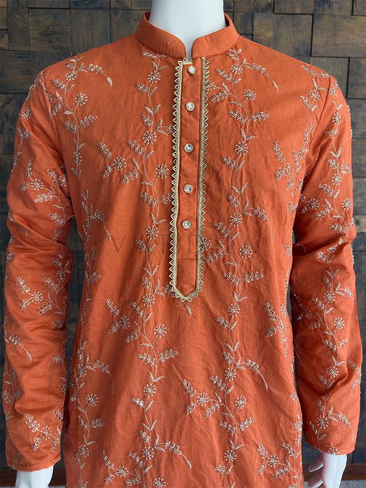 Orange kurta with intricate gold embroidery on a wooden background. Side profile of the terracotta orange embroidered viscose kurta for men, highlighting the luxurious drape. This fabric provides a luxurious, silk-like drape and a beautiful natural sheen. It flows elegantly over the body and carries the all-over embroidery perfectly without feeling stiff or heavy.