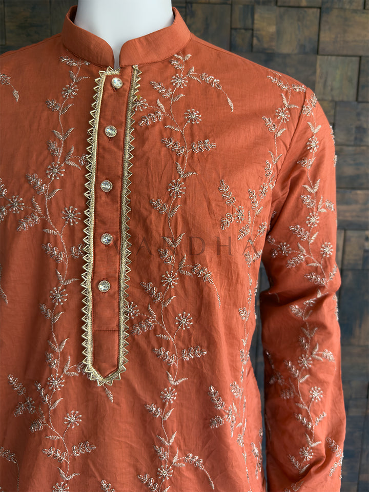 Orange embroidered kurta on a mannequin against a dark textured wall. Close-up of the full sleeve showing detailed floral embroidery on the vibrant orange viscose fabric. Unlike the previous minimalist pieces, this garment features exquisite, all-over delicate floral and vine embroidery. The threadwork catches the light beautifully. The center placket is highly detailed, framed by a striking gold zig-zag (chevron) zari border, creating a sharp focal point amidst the floral pattern.