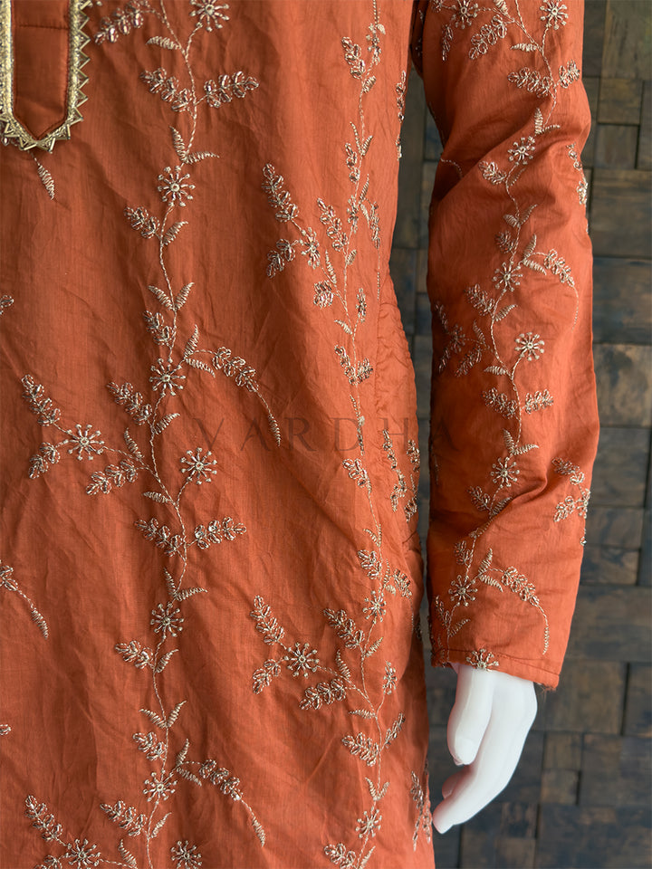 Orange embroidered garment on a mannequin against a brick wall. This is a vibrant, celebratory Terracotta Orange Rust long kurta. The bright, warm hue paired with heavy all-over work makes this an absolute showstopper, perfectly targeted for Haldi ceremonies, Sangeet nights, and grand festive occasions.