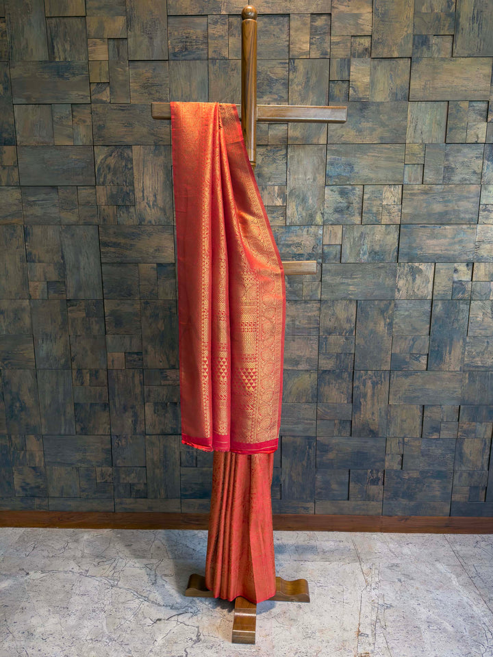 Red saree with gold patterns on a wooden stand against a textured wall. Vermilion Red Kanjivaram Silk Saree with golden floral zari motifs