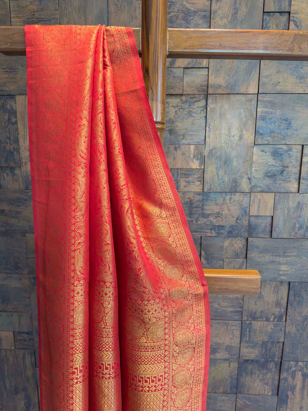 Red fabric with gold patterns draped over a wooden rod against a blue tiled wall. Close-up of broad red border featuring circular and geometric gold zari patterns