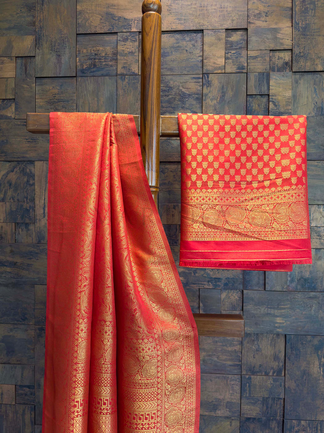 Red saree with gold patterns draped over a wooden rod against a textured blue wall. Close shot of gold zari craftsmanship on red saree body and border