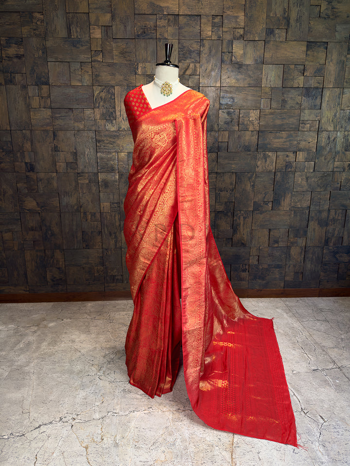 Red saree with gold patterns on a mannequin against a textured wall. House of Vardha premium deep pinkish-red silk saree showcasing all-over gold zari brocade work draped on a mannequin.