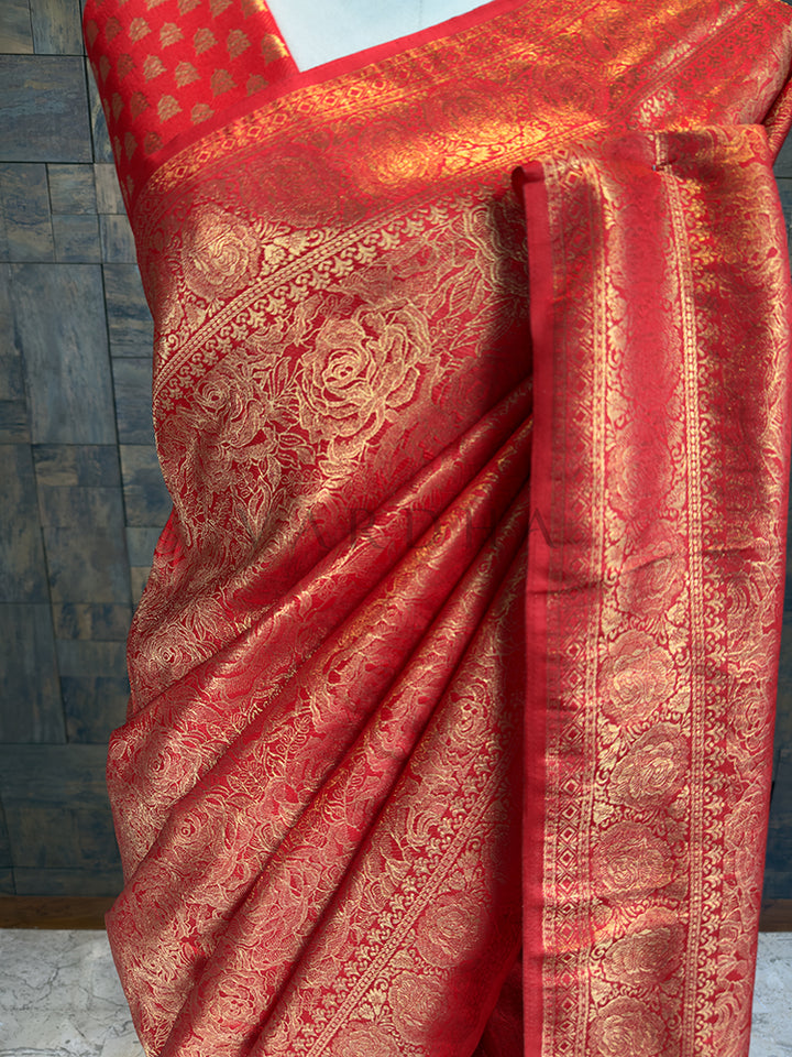 Close-up of House of Vardha rich magenta-red silk saree highlighting detailed gold zari craftsmanship on the pleats and body. Red saree with gold patterns draped over a surface