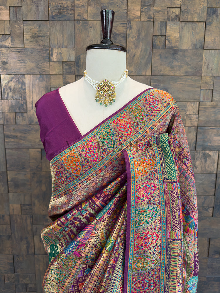 Colorful saree with intricate patterns on a mannequin against a wooden paneled wall. Close-up of a royal purple traditional silk saree draped over the shoulder, highlighting vibrant woven floral, geometric, and traditional motifs in multi-color Meenakari threadwork, accessorized with a classic pearl and kundan choker.