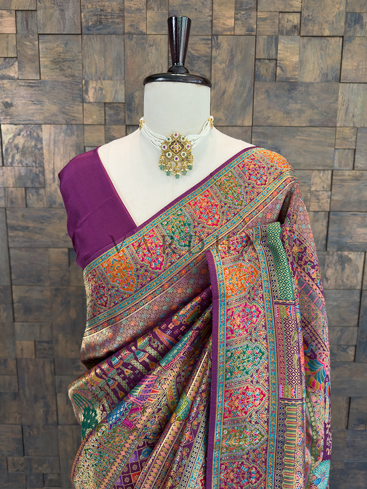Colorful saree with intricate patterns on a mannequin against a wooden paneled wall. Close-up of a royal purple traditional silk saree draped over the shoulder, highlighting vibrant woven floral, geometric, and traditional motifs in multi-color Meenakari threadwork, accessorized with a classic pearl and kundan choker.