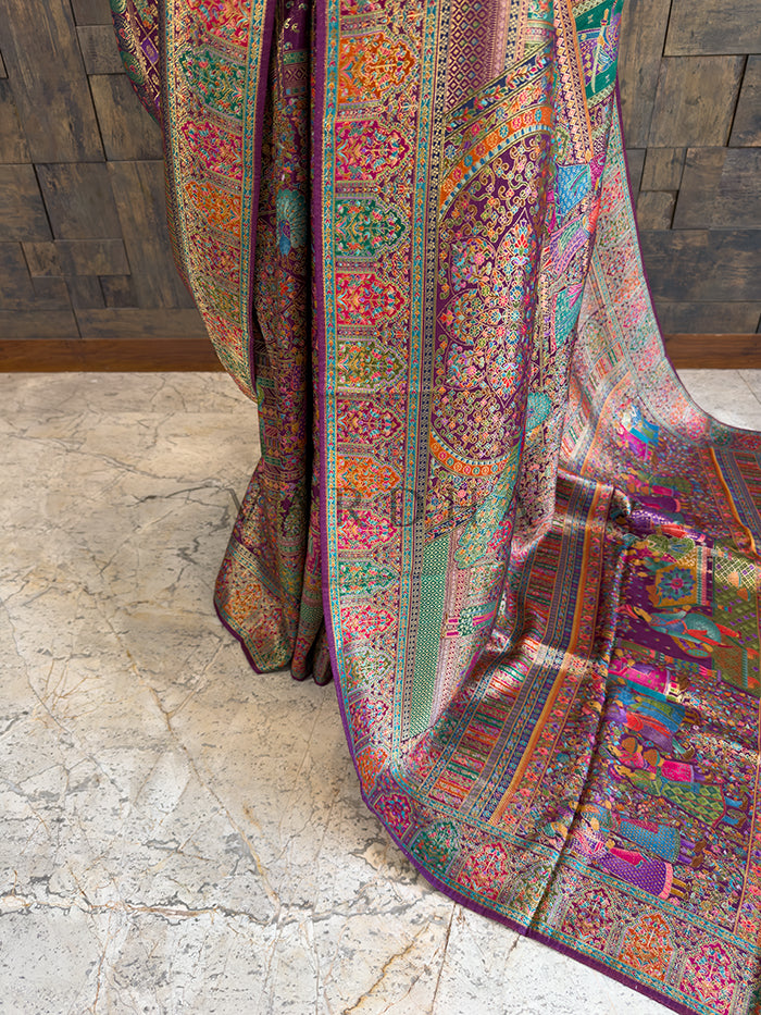 Colorful patterned fabric draped over a wooden surface Lower drape of a heavy heritage woven Meenakari silk saree, showcasing the rich purple base and elaborate multi-colored borders with woven floral vines and traditional human figure motifs cascading elegantly toward the floor.