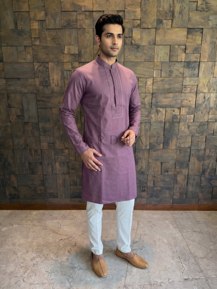 Man wearing a purple kurta with white pants against a wooden paneled wall. A rich Wine-colored cotton kurta for men, featuring a structured chest panel with central vertical pintucks and a crisp Mandarin collar.