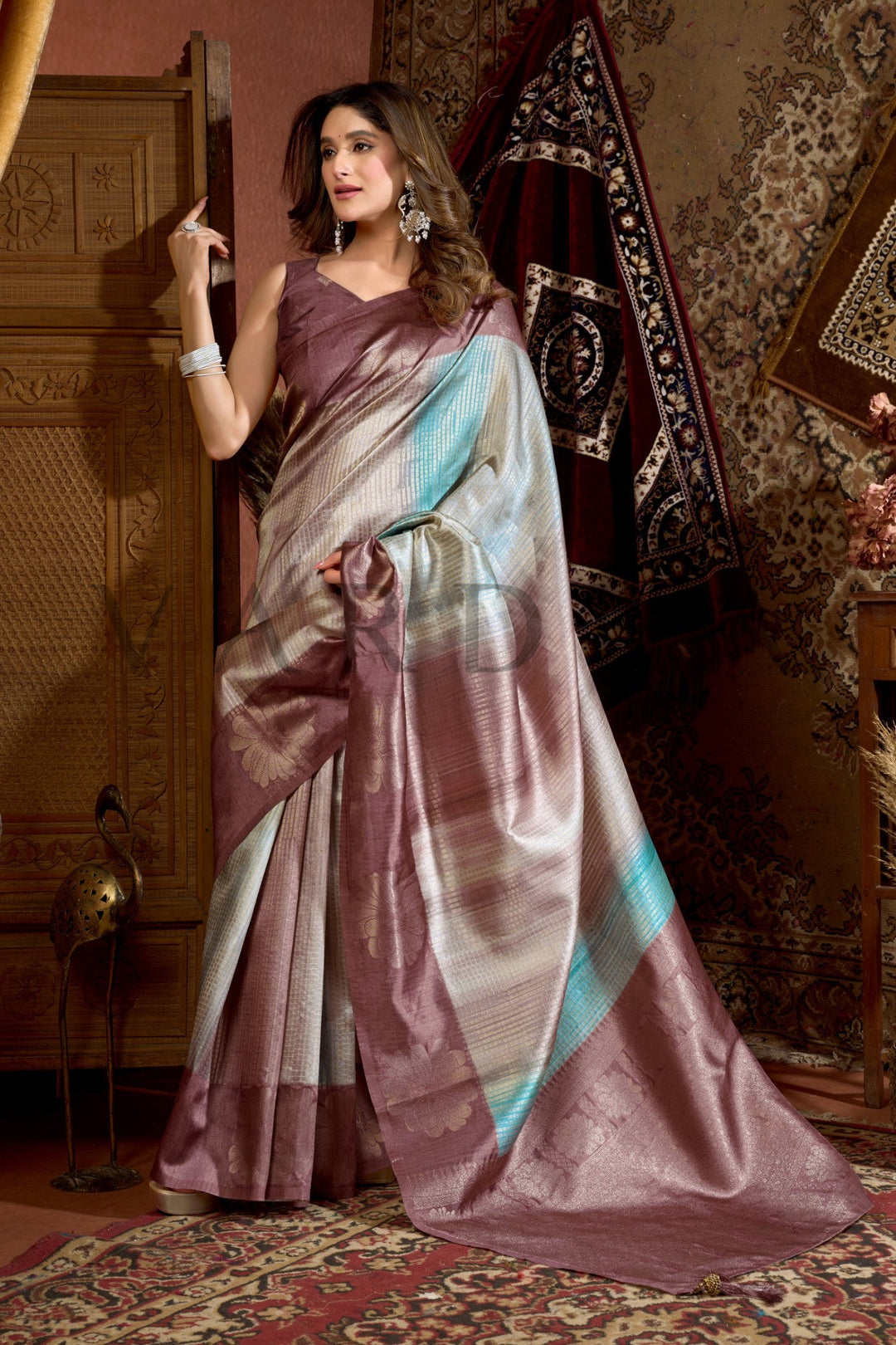 Woman wearing a traditional saree in a decorative room with patterned walls and rug. Enjoy premium add-ons including Pre-drape Services, Custom Stitched Blouse at a Complimentary Price, Fall and Edging, Gift Packaging, and Free Shipping!