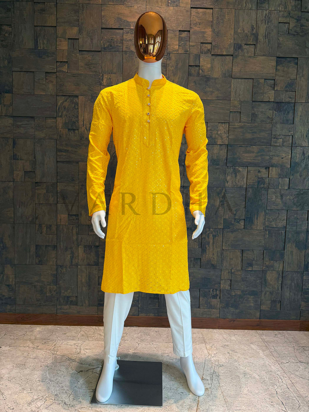 Mannequin wearing a yellow kurta against a textured wall. Sunflower yellow viscose men’s kurta with mandarin collar, ornate buttons and jacquard lattice sprinkled with sequins, displayed on mannequin with white churidar for festive wear.