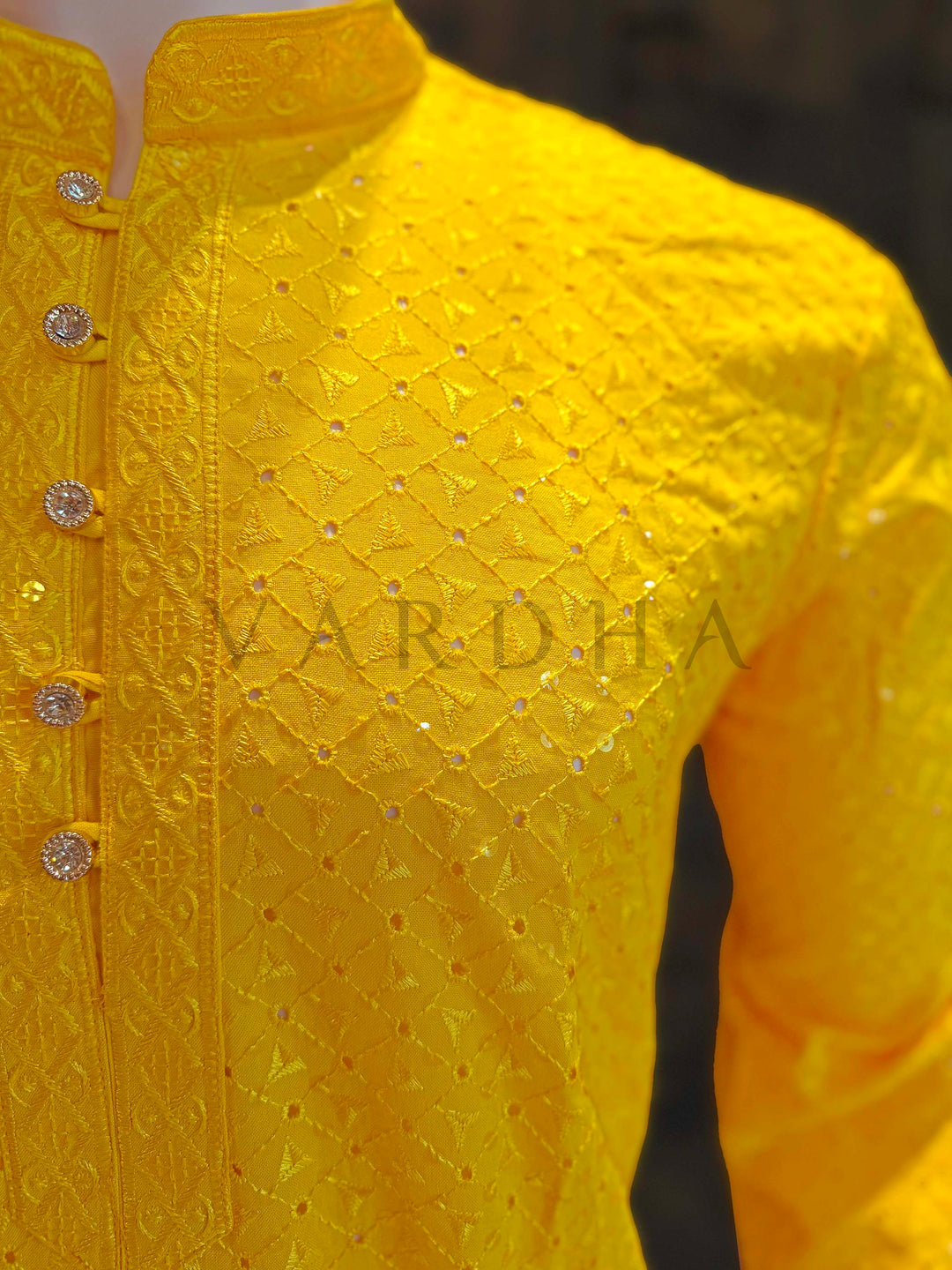 Yellow embroidered garment with 'Vardha' branding on a blurred background Angled shot emphasizing crisp mandarin collar, embellished buttons and evenly distributed sequin details on vibrant yellow festive kurta.