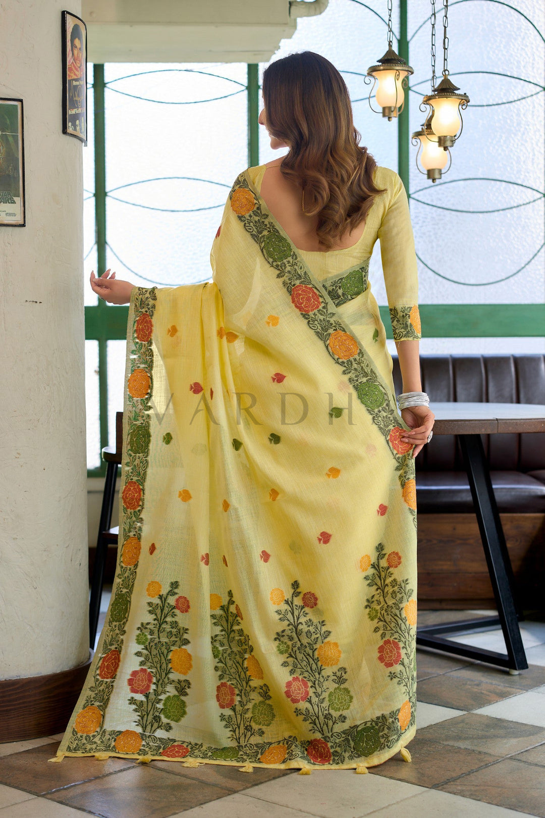 Woman wearing a yellow saree with floral patterns indoors. Back view of the yellow linen jacquard saree displaying the stunning oversized woven floral vines and matching yellow fancy tassels on the designer pallu.