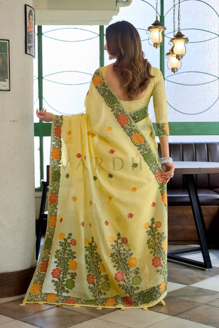 Woman wearing a yellow saree with floral patterns indoors. Back view of the yellow linen jacquard saree displaying the stunning oversized woven floral vines and matching yellow fancy tassels on the designer pallu.