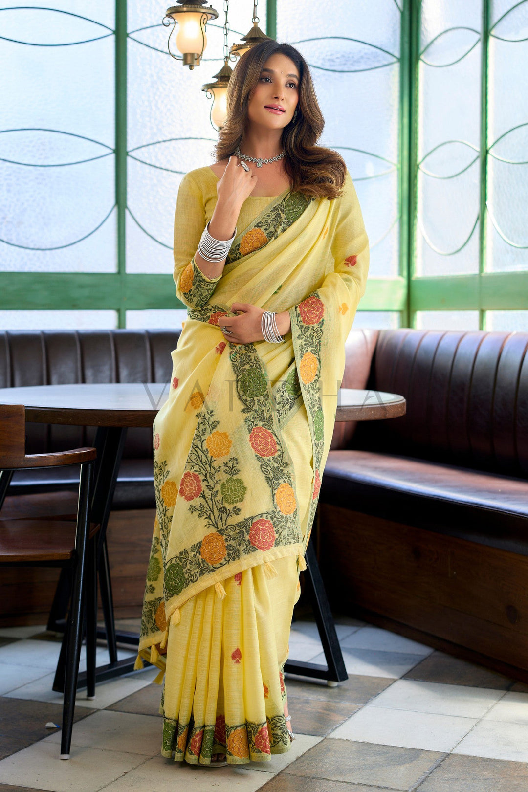 Woman in a yellow saree with floral patterns in an indoor setting Model leaning elegantly, displaying the lightweight and breathable drape of the yellow linen handloom saree, perfect for daytime festive celebrations and pujas.