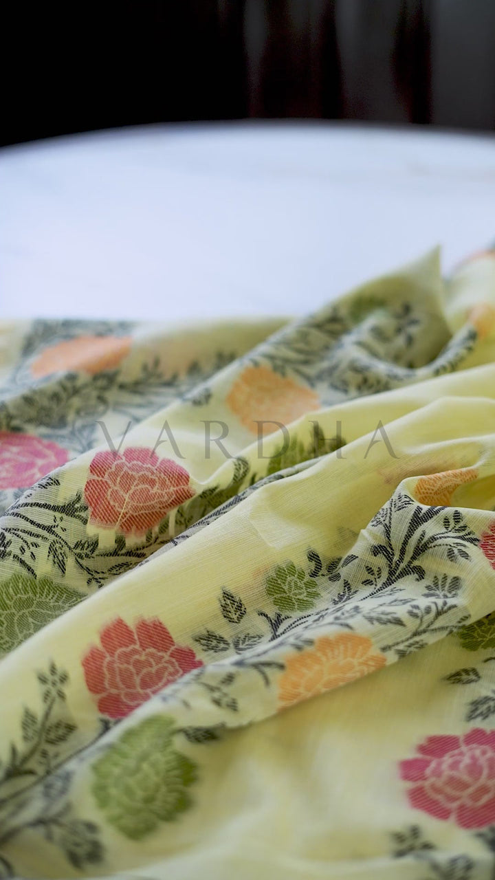 A close-up of a light yellow fabric with a colorful floral block print pattern, draped elegantly. The fabric features pink, orange, and green flowers with dark green leafy stems. Close-up macro detail of the yellow linen saree fabric showcasing the intricate multicolored woven floral border, scattered body motifs, and breathable handloom texture.