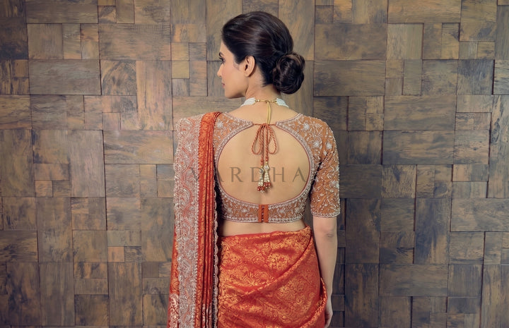 Woman wearing a traditional red saree with intricate designs against a wooden paneled wall.
