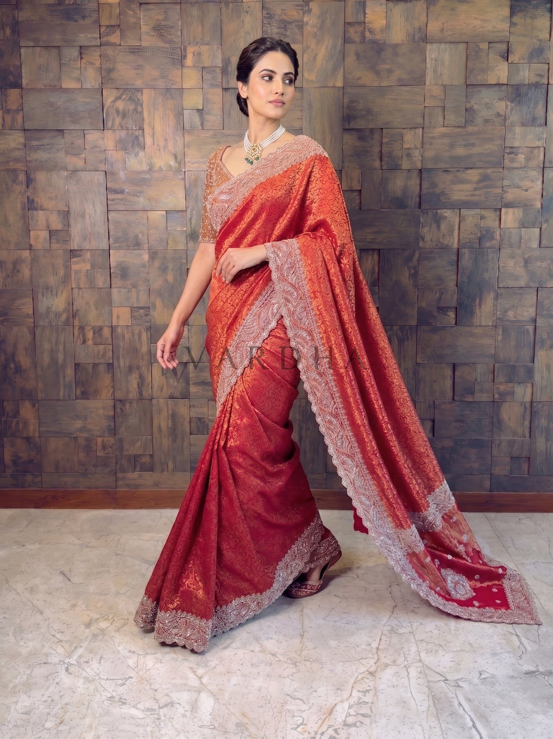 Woman wearing a red saree with gold patterns against a textured wall.